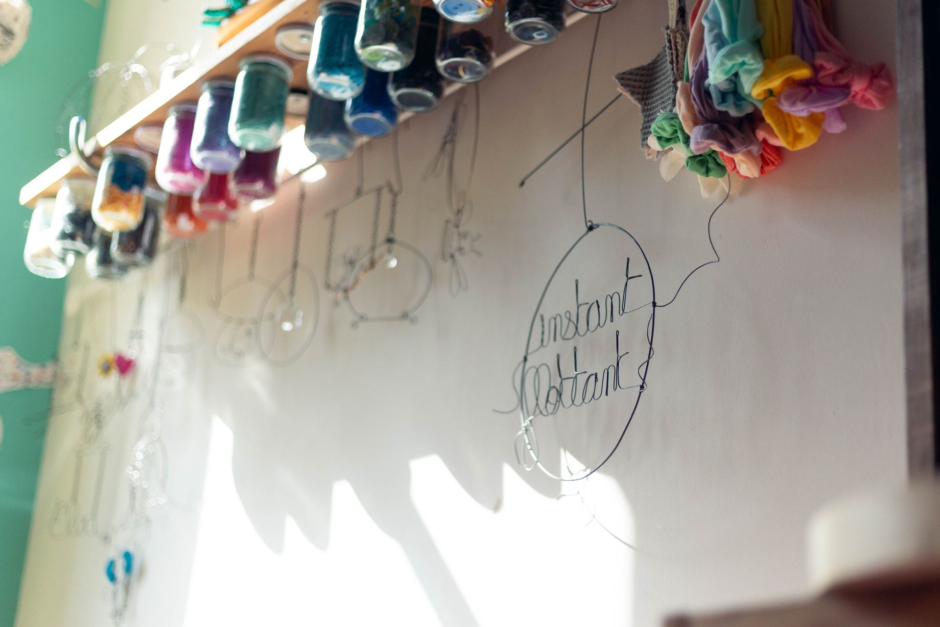 Photographie d'une enseigne en fil de fer avec l'inscription cursive « Instant Flottant » suspendue dans l'atelier avec les fils de laines colorés au dessus